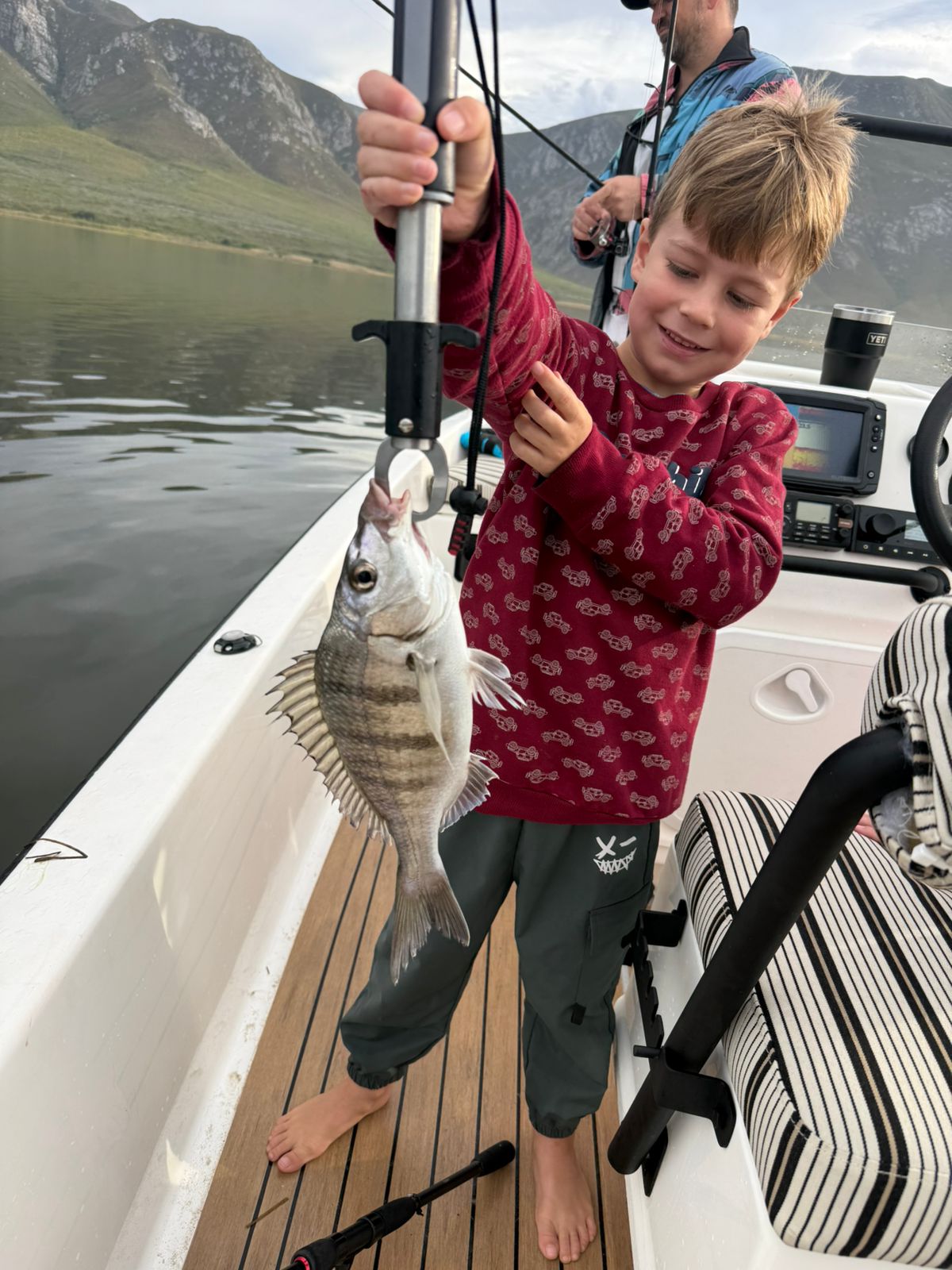 Boy on a boat holding up a freshly caught fish with the mountain behind